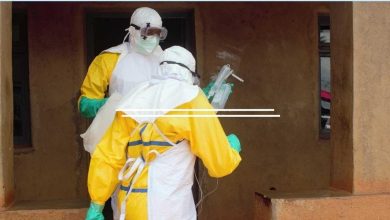 Two healthcare workers in full PPE (yellow suits, masks, face shields) prepare an IV bag outside a tan building.