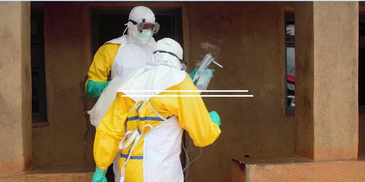 Two healthcare workers in full PPE (yellow suits, masks, face shields) prepare an IV bag outside a tan building.
