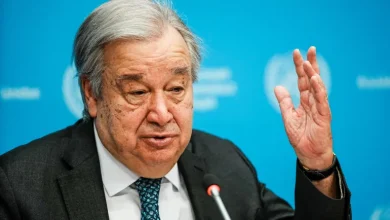 Older man in a dark suit speaks at a press conference, raising his right hand as a microphone is in front of him against a blue backdrop.