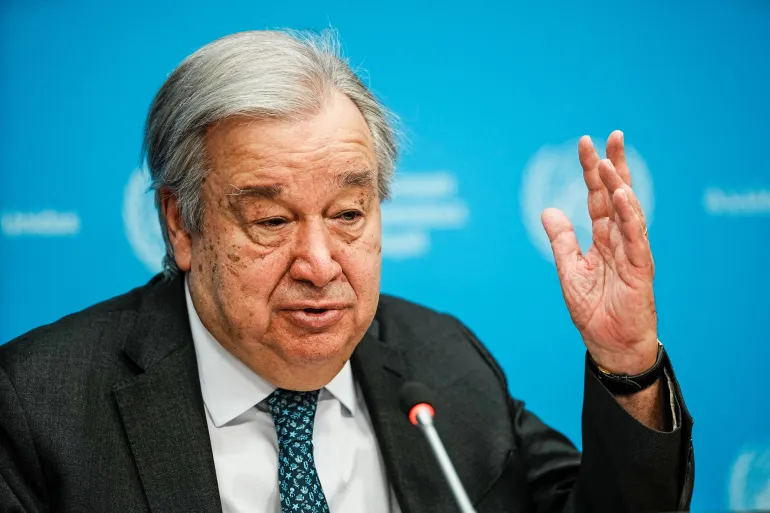 Older man in a dark suit speaks at a press conference, raising his right hand as a microphone is in front of him against a blue backdrop.