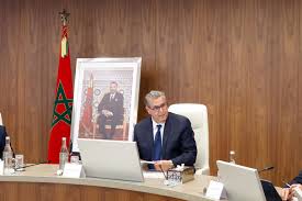 images 20 | MCG24 | موقع إلكتروني مغربي شامل متجدد على مدار الساعة Official in a suit seated at a wooden desk with two laptops, Moroccan flag behind him and a framed portrait on the wall.