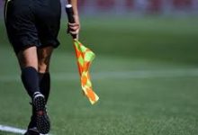 Assistant referee walking on a soccer field, holding an orange checkered flag.