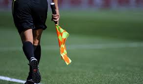 Assistant referee walking on a soccer field, holding an orange checkered flag.
