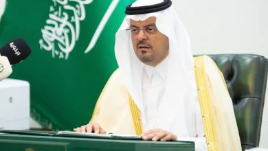 Saudi official in traditional white attire sits at a green desk with a microphone and keyboard, Saudi flag in the background