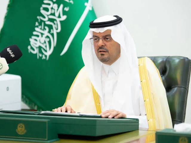 Saudi official in traditional white attire sits at a green desk with a microphone and keyboard, Saudi flag in the background