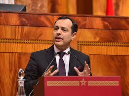 Man in a dark suit speaks at a red podium in a formal chamber with wood paneling behind him.
