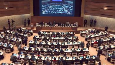 Large international conference hall with delegates seated at curved desks facing a central stage and big screen projection above the podiums