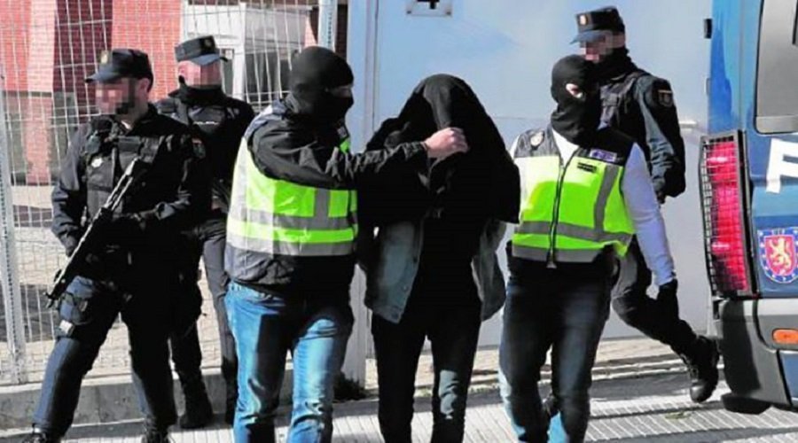 Masked police officers escort a hooded detainee toward a blue van during an arrest on a street edge with a visible reflective vest.