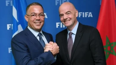 Two men in suits shake hands in front of a FIFA backdrop, with a Moroccan flag visible on the right.