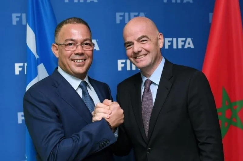 Two men in suits shake hands in front of a FIFA backdrop, with a Moroccan flag visible on the right.