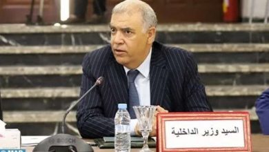 Man in a pinstripe suit speaks at a formal meeting; on the table is a sign in Arabic reading 'السيد وزير الداخلية' (Mr. Minister of Interior).