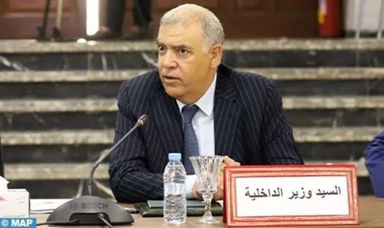 Man in a pinstripe suit speaks at a formal meeting; on the table is a sign in Arabic reading 'السيد وزير الداخلية' (Mr. Minister of Interior).