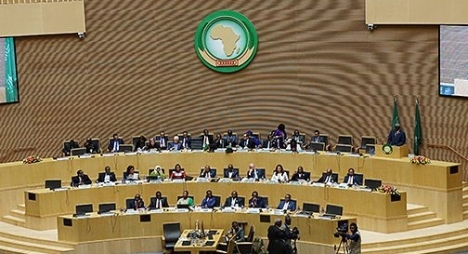 Africa Union emblem above a tiered semicircular council chamber where delegates sit around long desks in formal session.