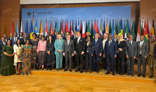 Large group of dignitaries and international delegates posing on a stage with rows of national flags in the background, during a formal ceremony.