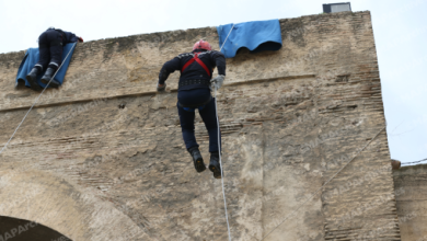 Person rappelling down a large stone wall with a safety rope, while another climber sits near the top under blue tarps.