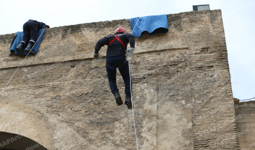 Person rappelling down a large stone wall with a safety rope, while another climber sits near the top under blue tarps.