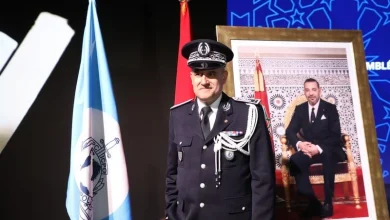 maroc-interpol-2025 | MCG24 | موقع إلكتروني مغربي شامل متجدد على مدار الساعة Uniformed officer in dress uniform stands beside a blue flag and a red flag, with a large framed portrait of a seated man in the background.