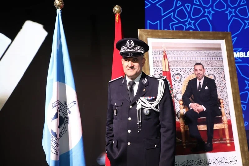 maroc-interpol-2025 | MCG24 | موقع إلكتروني مغربي شامل متجدد على مدار الساعة Uniformed officer in dress uniform stands beside a blue flag and a red flag, with a large framed portrait of a seated man in the background.