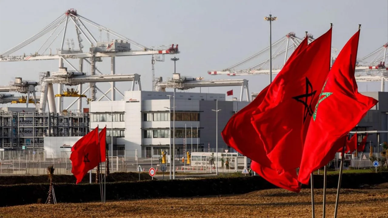 Red Moroccan flags wave in the foreground at a port, with large cranes and a modern building in the background.