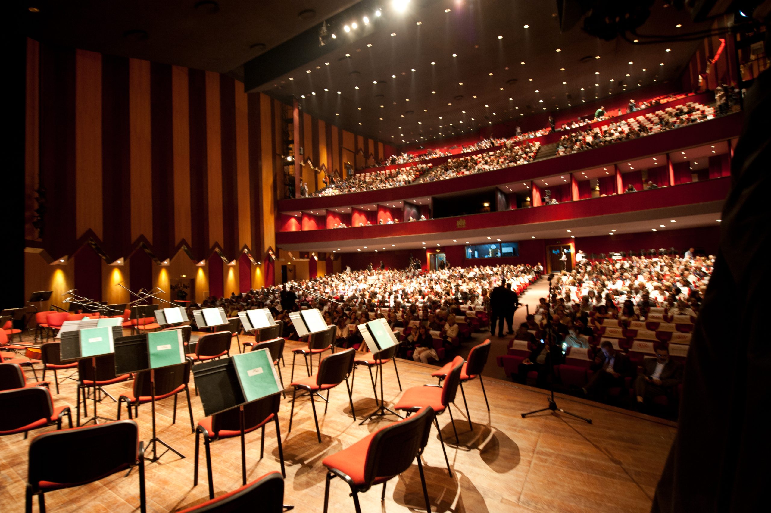Concert hall interior with a stage full of empty chairs and music stands in the foreground and a large audience seated in red seats on multiple balconies above.