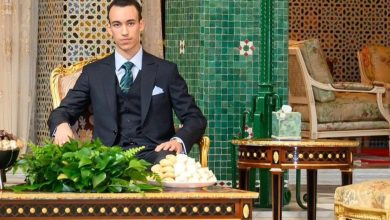 Young man in a dark suit sits at a carved table with green foliage and pastries in a luxurious, tiled room.