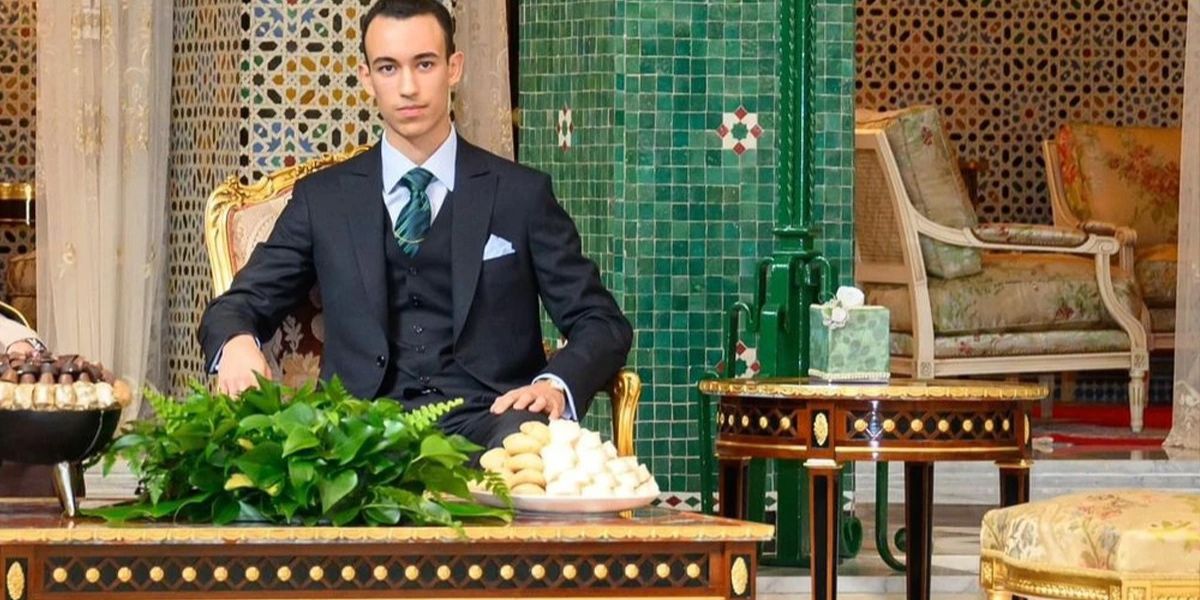 Young man in a dark suit sits at a carved table with green foliage and pastries in a luxurious, tiled room.