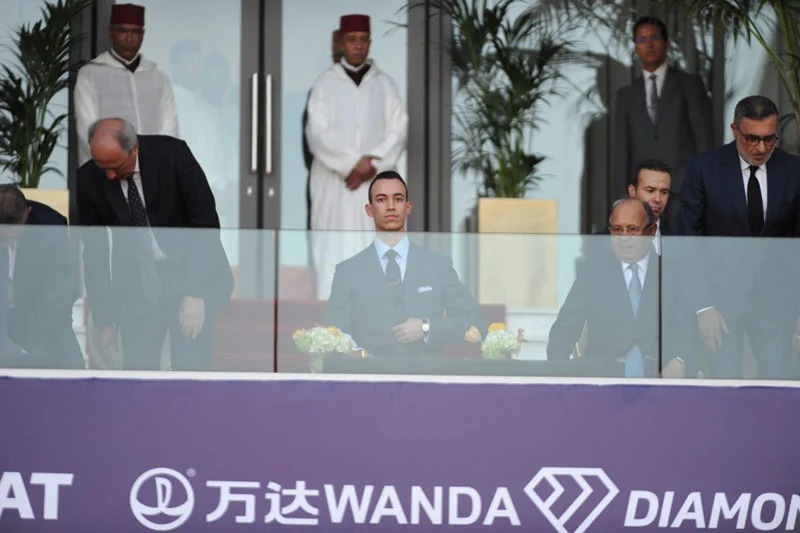 Man in a dark suit seated at a glass barrier during a formal event, with others standing behind, in front of a purple banner reading Wanda Diamonds.
