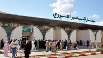 Group of travelers queueing outside a modern international airport terminal with curved white arches and a DEPART sign above the entrance.