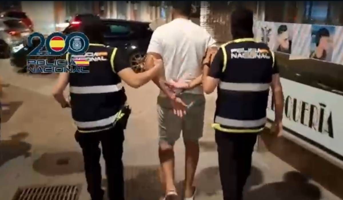 Two police officers walk with a handcuffed man down a city sidewalk at night, one on each side, guiding him toward a police vehicle for transport.
