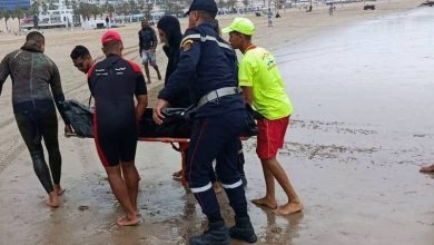 naufrage-1 | MCG24 | موقع إلكتروني مغربي شامل متجدد على مدار الساعة Emergency responders carry a person on a stretcher along the wet beach while others assist nearby workers and onlookers