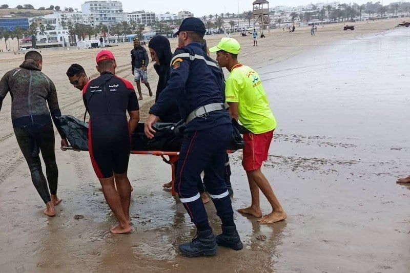 Emergency responders carry a person on a stretcher along the wet beach while others assist nearby workers and onlookers