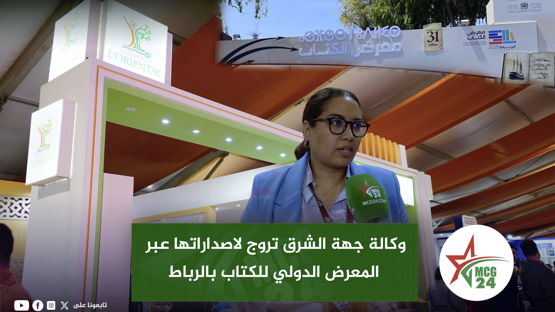 Woman with glasses speaking into a green microphone at a booth at a book fair; MCG24 logo visible, Arabic text overlay on a green banner in front of her.