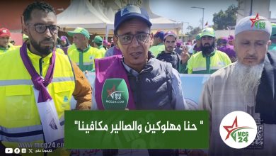 A man with glasses speaks into a green MCG24 microphone at a crowded outdoor event, with volunteers in bright jackets behind him and Arabic text overlay in front.