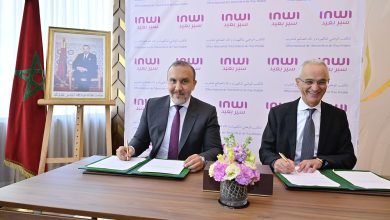 Two men in suits signing documents at a desk during a formal ceremony, with a Moroccan flag and a framed portrait to the left and a pink INWI backdrop behind them.