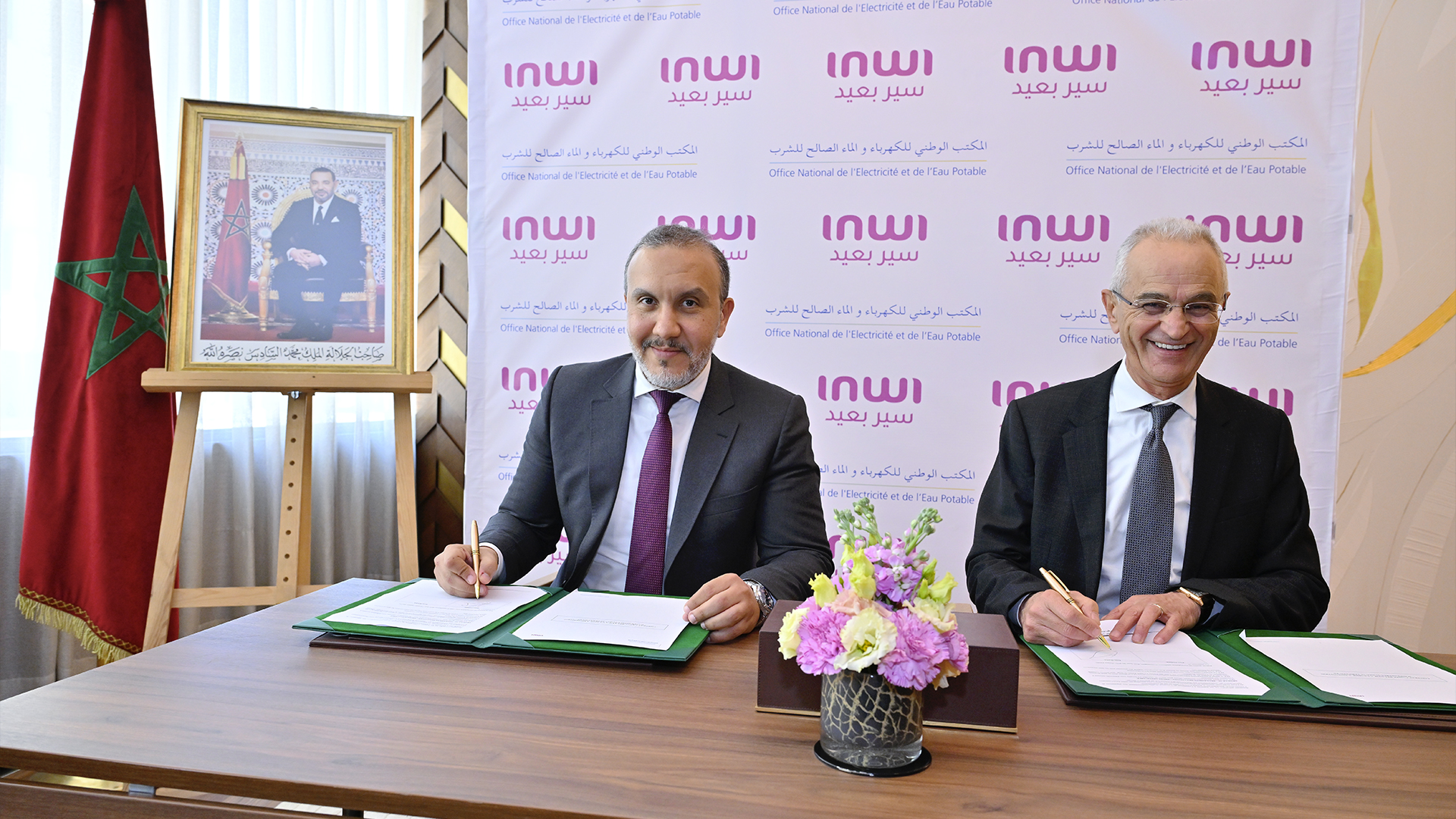 Two men in suits signing documents at a desk during a formal ceremony, with a Moroccan flag and a framed portrait to the left and a pink INWI backdrop behind them.