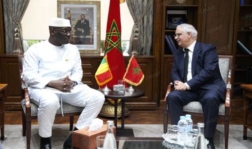 Two men in a formal office sit facing each other and discuss, with Moroccan flags between them and a framed portrait behind.