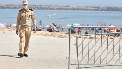 plage-800-2 | MCG24 | موقع إلكتروني مغربي شامل متجدد على مدار الساعة Beige-uniformed security guard walking along a sandy beach near a metal crowd-control barrier, with beachgoers in the background.