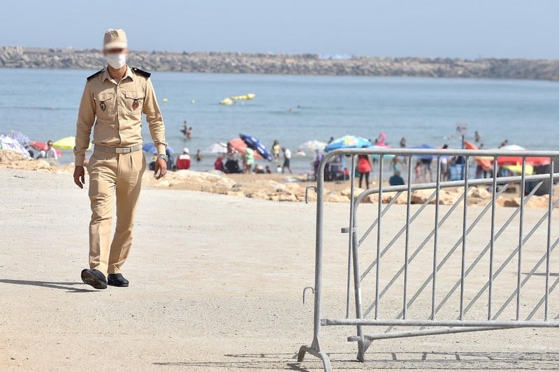 plage-800-2 | MCG24 | موقع إلكتروني مغربي شامل متجدد على مدار الساعة Beige-uniformed security guard walking along a sandy beach near a metal crowd-control barrier, with beachgoers in the background.