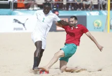 Two soccer players compete for the ball on a sandy beach, one in white (number 2) and one in red, with spectators in the background.