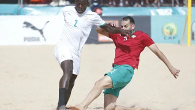 plpllp | MCG24 | موقع إلكتروني مغربي شامل متجدد على مدار الساعة Two soccer players compete for the ball on a sandy beach, one in white (number 2) and one in red, with spectators in the background.
