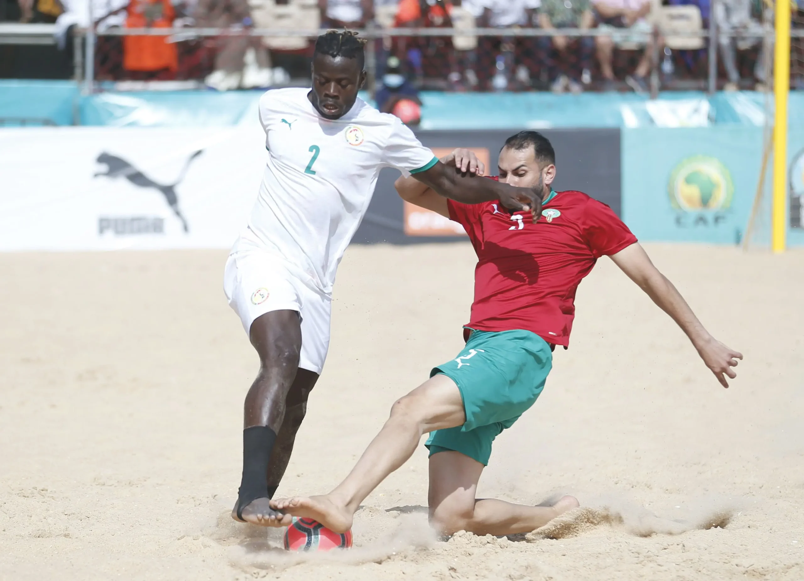 plpllp | MCG24 | موقع إلكتروني مغربي شامل متجدد على مدار الساعة Two soccer players compete for the ball on a sandy beach, one in white (number 2) and one in red, with spectators in the background.