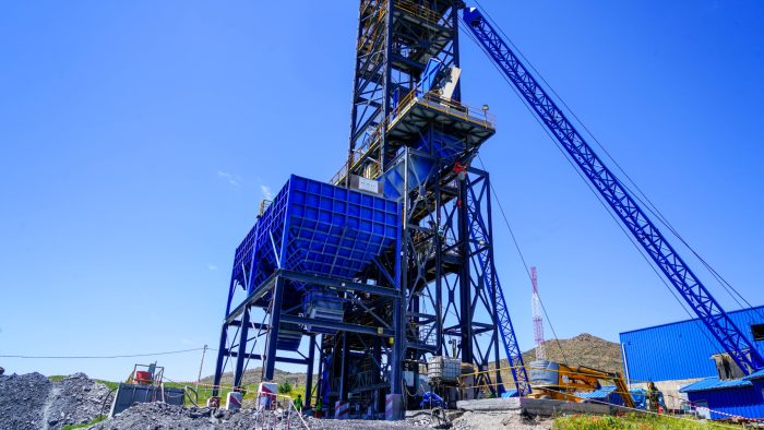 Heavy mining crane with a blue hopper elevated on a steel framework at a quarry site, with a long conveyor arm nearby