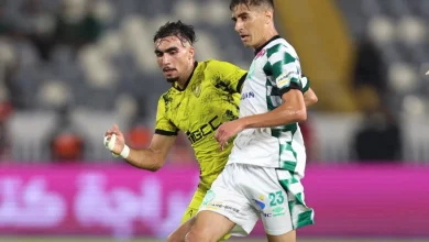 Two soccer players contest the ball during a match, one in a yellow kit and the other in a white-and-green striped kit, running side by side on the field.