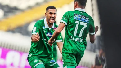 Two soccer players in green kits celebrate on the field, one facing forward with a fist raised in triumph.