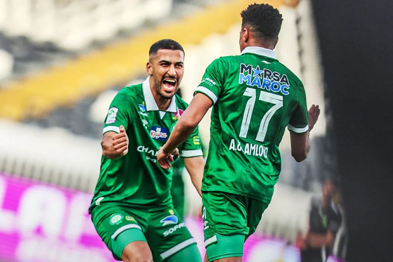 Two soccer players in green kits celebrate on the field, one facing forward with a fist raised in triumph.