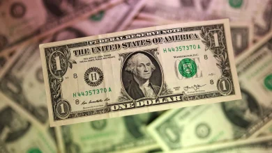 Close-up of a US one-dollar bill with George Washington's portrait, green serial numbers, and intricate border details as other bills blur in the background.