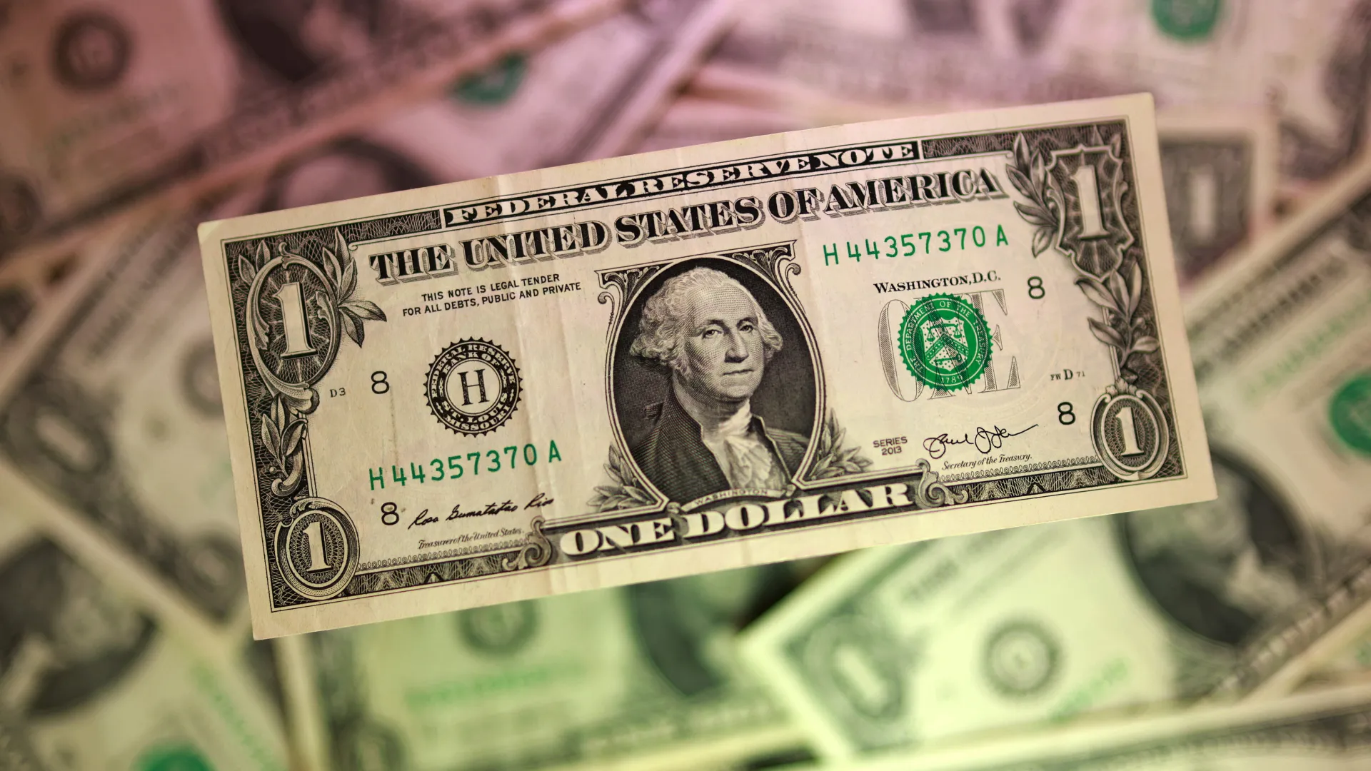 Close-up of a US one-dollar bill with George Washington's portrait, green serial numbers, and intricate border details as other bills blur in the background.