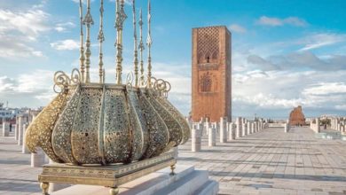 Ornate metal crown sculpture on a pedestal in a wide, tiled plaza, with a tall brick tower in the background.