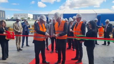Group of officials in hard hats and orange vests cut a red ribbon at an outdoor ribbon-cutting ceremony on a tarmac surface with a white tent in the background.