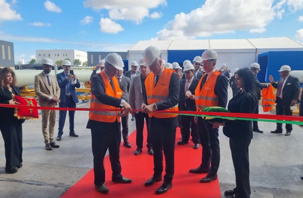 Group of officials in hard hats and orange vests cut a red ribbon at an outdoor ribbon-cutting ceremony on a tarmac surface with a white tent in the background.